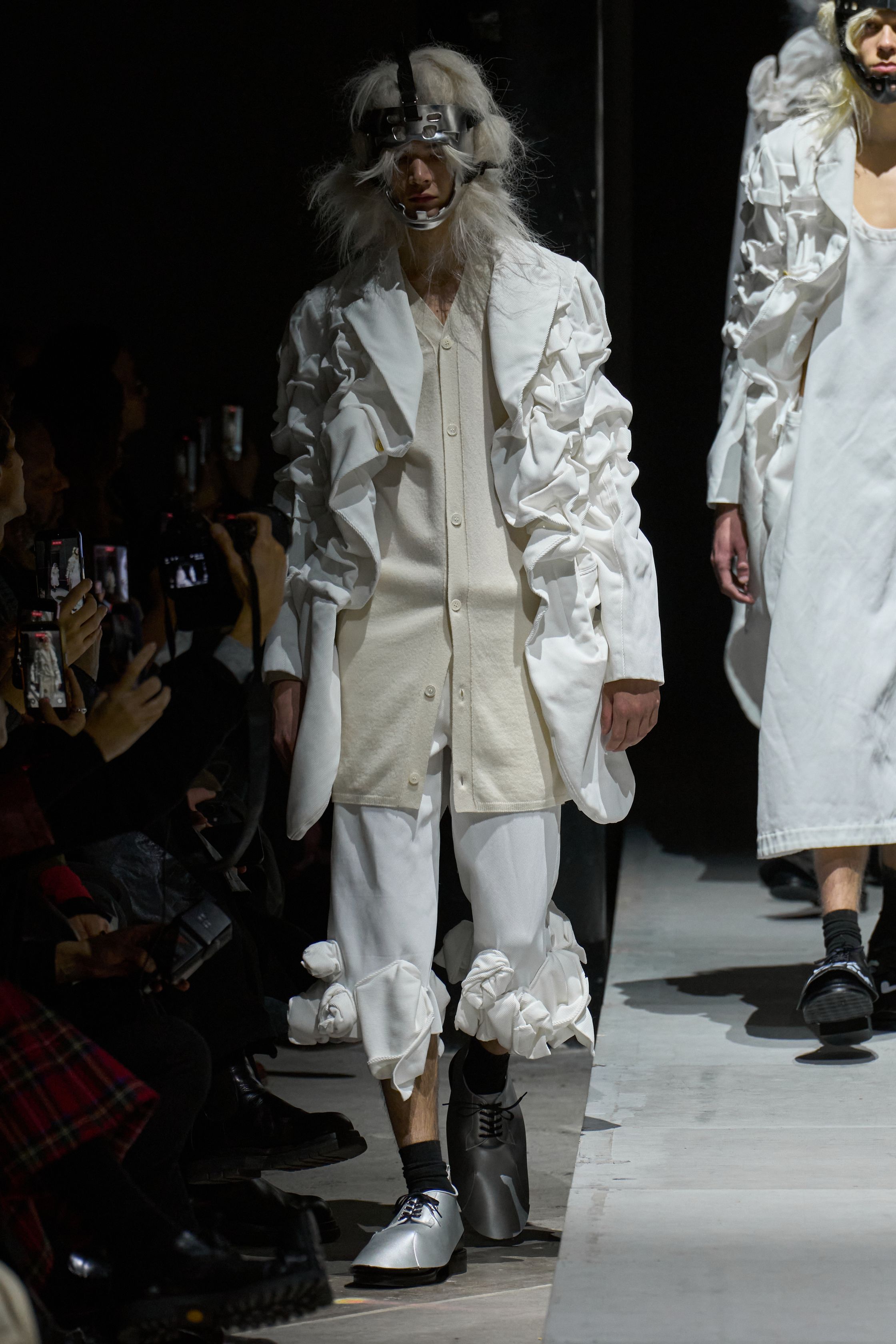 Comme des Garçons Homme Plus 2026秋冬以极端造型继续挑战“男性身体”的社会想象。面具、假发、解构西装与异化比例营造出近乎仪式化的压迫氛围，黑白灰成为思想载体而非装饰。这是一场关于权力、恐惧与集体意识的时装实验，服装不再服务于穿着，而是成为观念的武器-顽石频道趋势研究